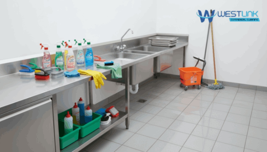 A clean commercial kitchen area with stainless steel counters, sinks, cleaning supplies, and a mop bucket, branded with the Westlink Commercial Cleaning