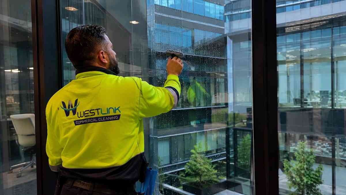 Westlink cleaner in a yellow safety jacket cleaning floor-to-ceiling windows inside a modern corporate office building.