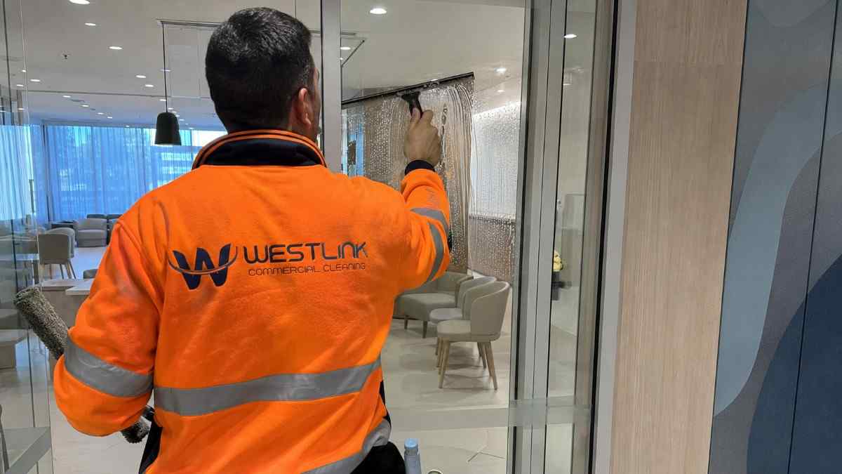 Westlink cleaner in an orange high-visibility jacket cleaning a glass partition inside a modern medical waiting area.