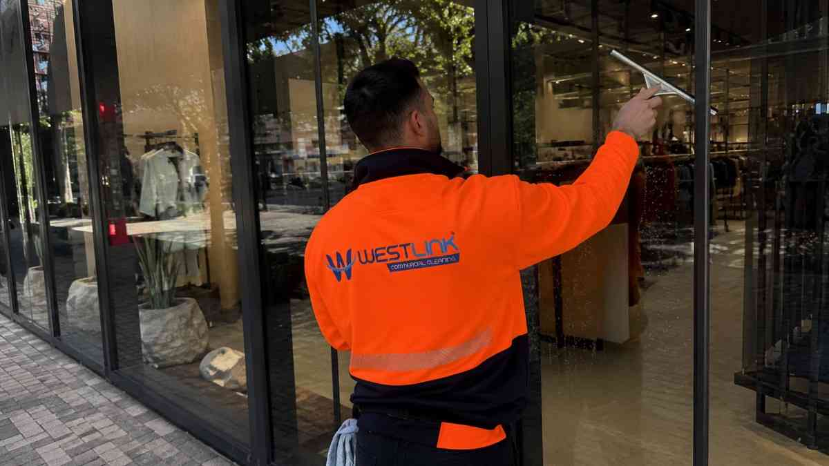 Westlink cleaner in an orange safety jacket using a squeegee on the exterior glass of a modern retail shopfront.