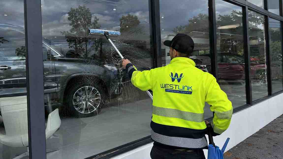 Westlink cleaner in a yellow safety jacket washing large dealership windows with vehicles visible inside the showroom.