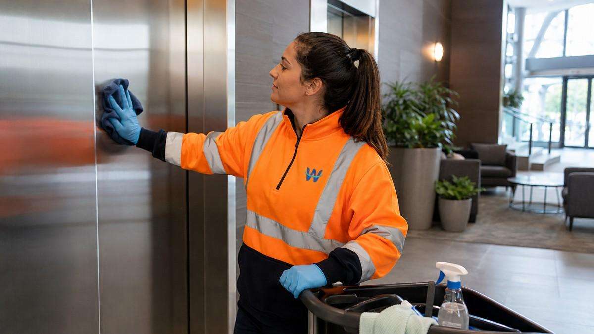 Westlink cleaner wiping stainless-steel elevator doors in a modern building lobby.