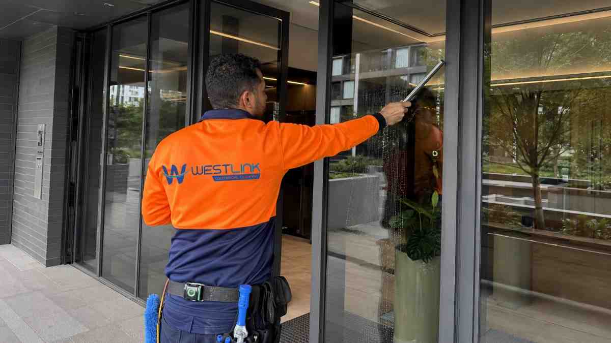 Westlink cleaner in an orange safety jacket cleaning the glass entrance of a modern strata apartment building.