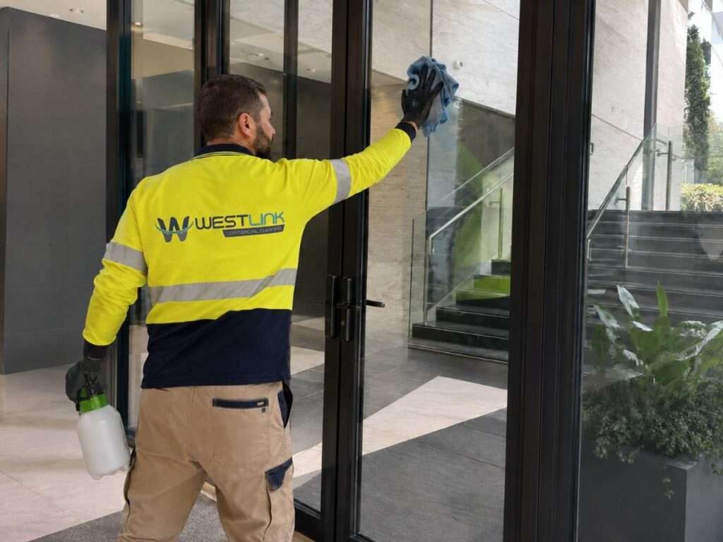 Westlink cleaner in high-visibility safety workwear wiping glass doors in a modern commercial building lobby.