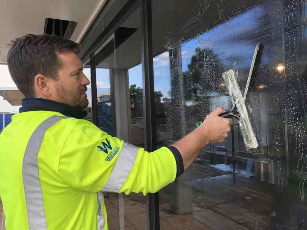 Westlink cleaner in a high-visibility yellow jacket washing a large commercial glass window with a squeegee.