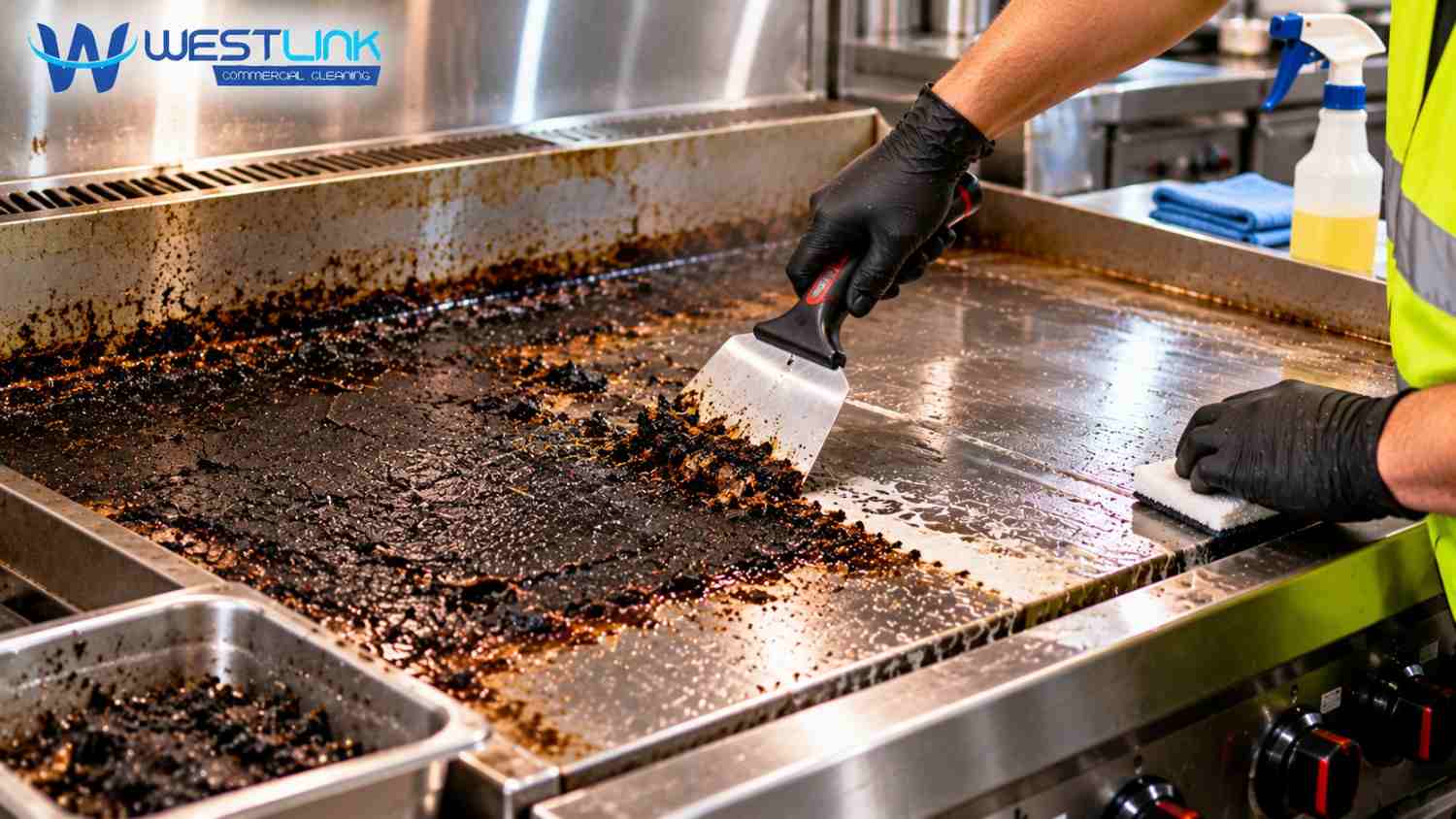 Commercial cleaner wearing protective gloves using a metal scraper to remove thick black carbon buildup from a flat-top griddle in a professional kitchen.